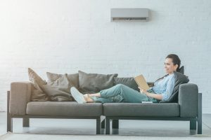 Happy,Woman,Reading,Book,On,Couch,,Air,Conditioner,On,Wall,